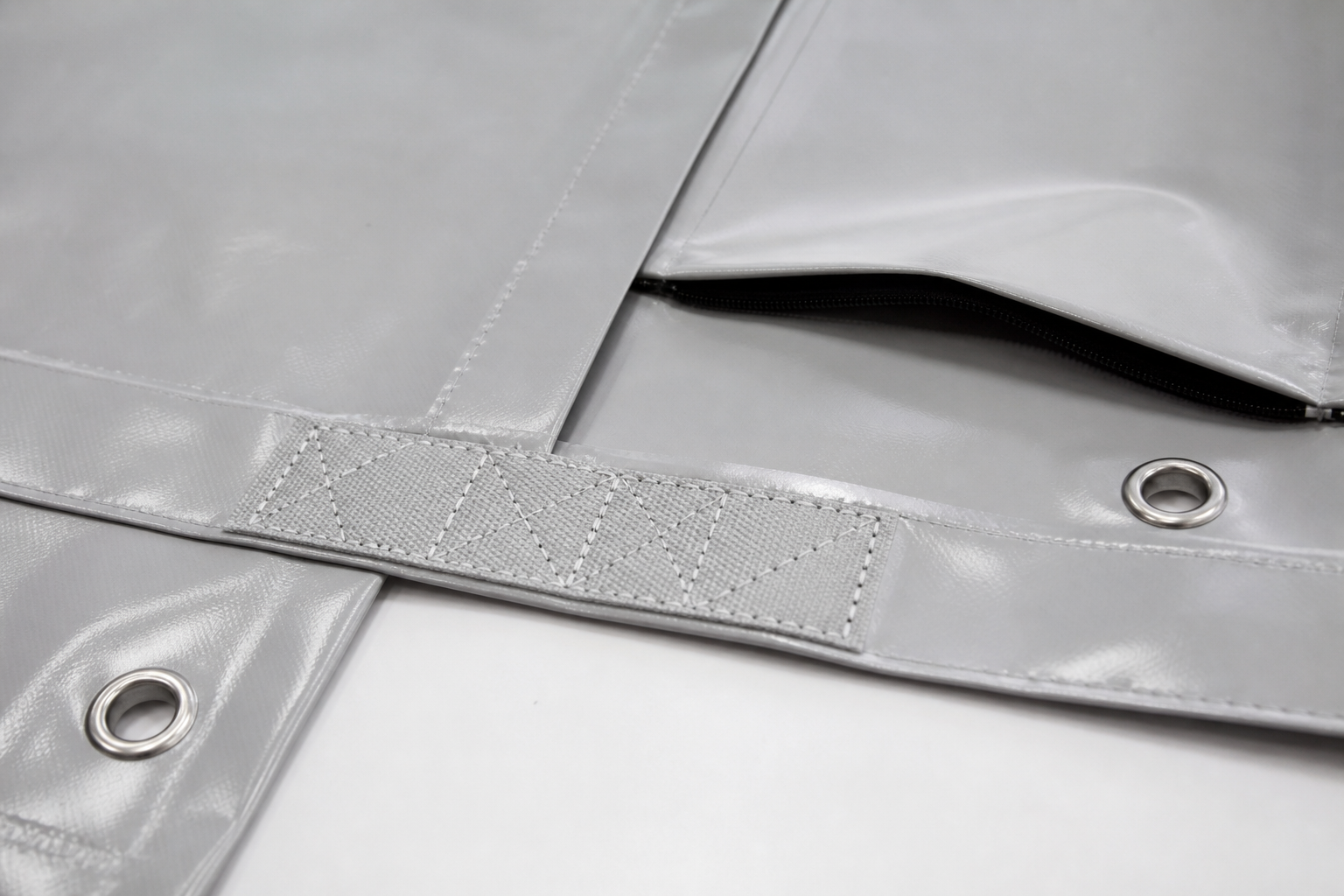 Close industrial detail of a custom PVC tarp with reinforced welded hem, metal eyelets, webbing, zipper access flap, and corner reinforcement.
