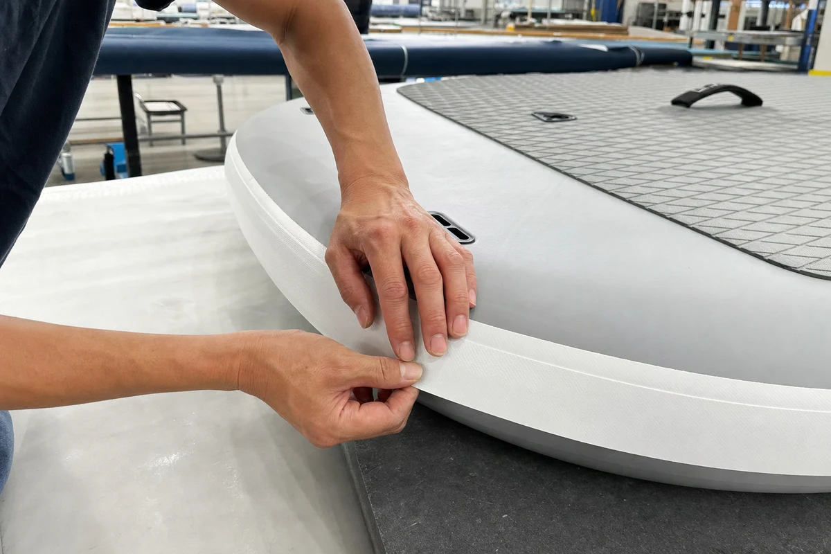 Factory technician aligning a thin PVC rail band around an inflatable paddle board perimeter